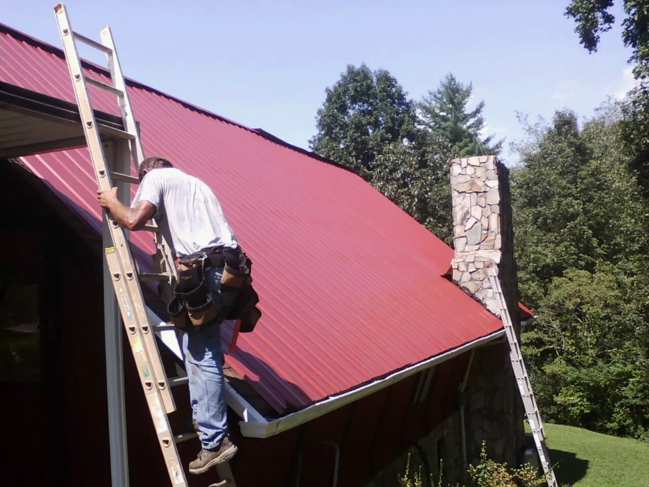 Roofer on a ladder beside a red metal roof and stone chimney — Soffit & Fascia Repair by Roofing Brothers in Lacota